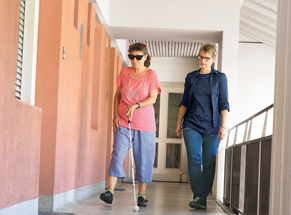 Mäde Müller hilft einer Klientin den Weg zu ihrer Freundin zu memorisieren. Die Klientin orientiert sich mit dem Langstock.